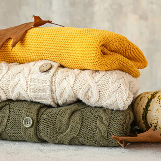 Stack of three knitted sweaters in yellow, white, and green with autumn leaves and pumpkins on a neutral background.