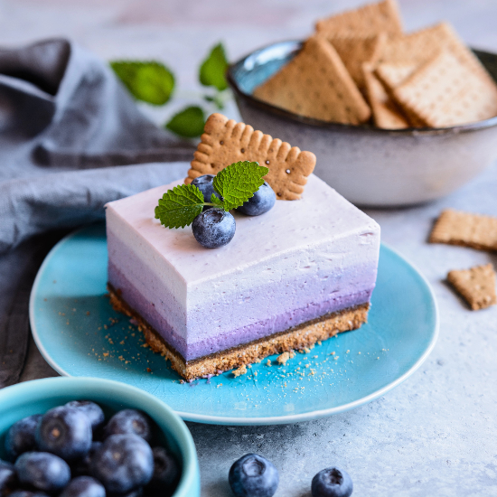 Slice of layered dessert with blueberries and a cookie on a blue plate, surrounded by more blueberries and cookies.