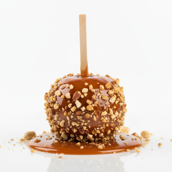 Caramel apple on a stick with nuts on a white background. the apple is set in a puddle of caramel.