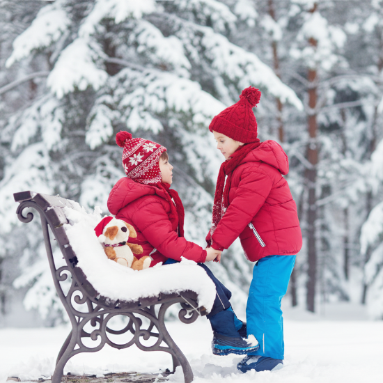 Two children in winter clothing sitting on a bench in a snowy forest.