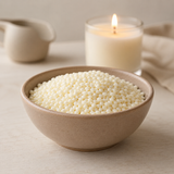 A beige ceramic bowl filled with wax beads, set on a light wooden surface with a lit cream candle and neutral home decor in the background.