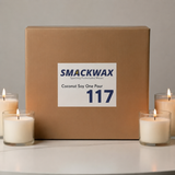 A brown cardboard box labeled SMACKWAX Coconut Soy One Pour 117 sits on a light surface with four lit white candles around it against a neutral gray background.