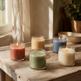 Four lit glass jar candles in assorted colors—rust red, pale yellow, sage green, and dusty blue—arranged on a rustic wooden table in a softly lit interior setting.