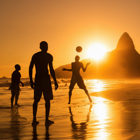 Silhouettes of people playing soccer on a beach at sunset.