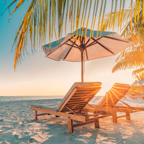Two lounge chairs under a sun umbrella on a sandy beach with palm trees and ocean view.