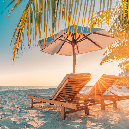 Two lounge chairs under a sun umbrella on a sandy beach with palm trees and ocean view.