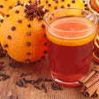 Glass of red beverage with orange slice, surrounded by oranges with star anise and cinnamon sticks on a wooden surface.