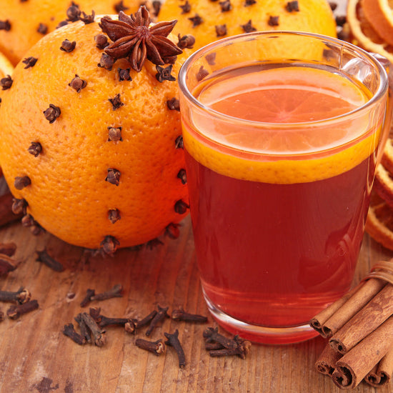 Glass of red beverage with orange slice, surrounded by oranges with star anise and cinnamon sticks on a wooden surface.