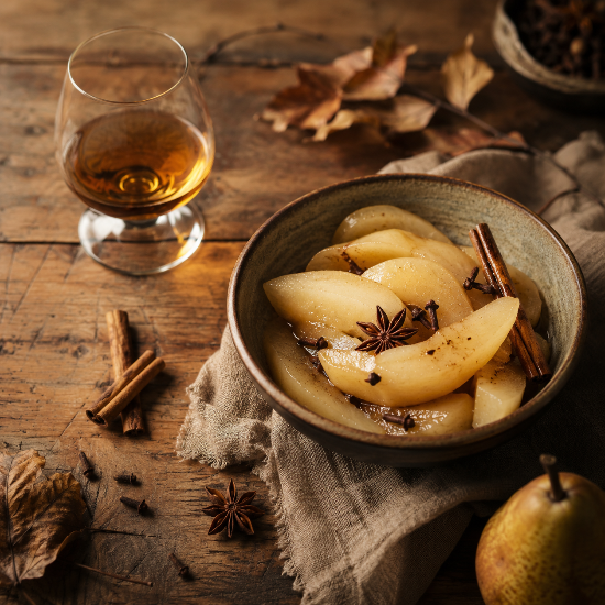 Bowl of poached pears with cinnamon and star anise on a wooden table with a glass of amber liquid.