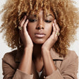 Woman with blonde curly hair wearing a brown jacket against a white background