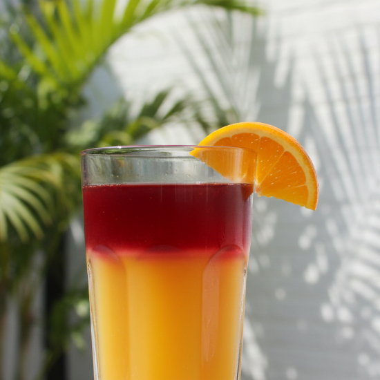 refreshing glass of mango juice topped with cherry juice and garnished with an orange wedge