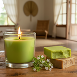 Green candle in a glass holder with two bars of soap and a small plant on a wooden surface.