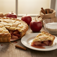 Apple pie with a slice on a plate, surrounded by apples and cinnamon sticks on a wooden table.