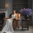 Woman in a white dress sitting in a brown leather armchair in a room with a fireplace and bookshelf.