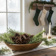 Decorative bowl with pinecones and greenery on a table, with stockings hanging in the background.