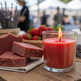Red candle in a glass jar with soap bars on a wooden table outdoors.