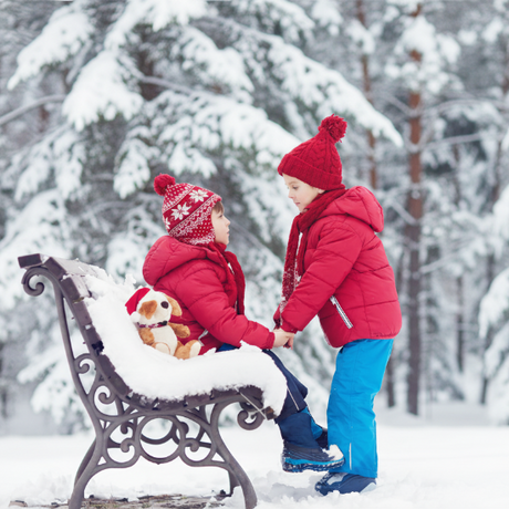 Two children in winter clothing sitting on a bench in a snowy forest.