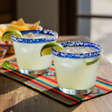 two margaritaa in Mexican style glasses with blue rim. there is white salt on the rim. garnished with a lime. bright and refreshing. 