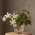 Clear glass vase with white flowers and green leaves on a wooden surface against a brown background
